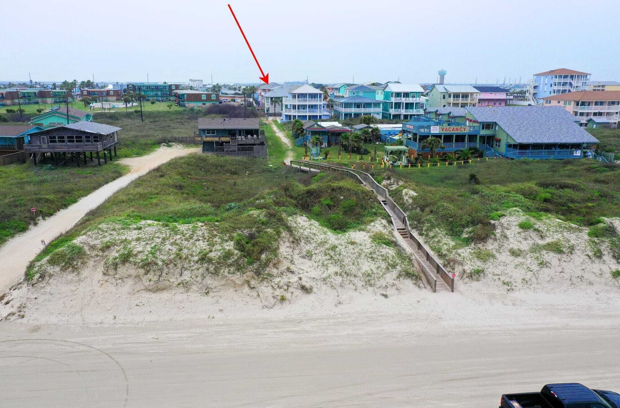 A beachfront property with multiple buildings, including a house named 'The Sunshine House' and a 'VACANCY' sign. There is a sandy beach and a wooden boardwalk leading to the buildings.
