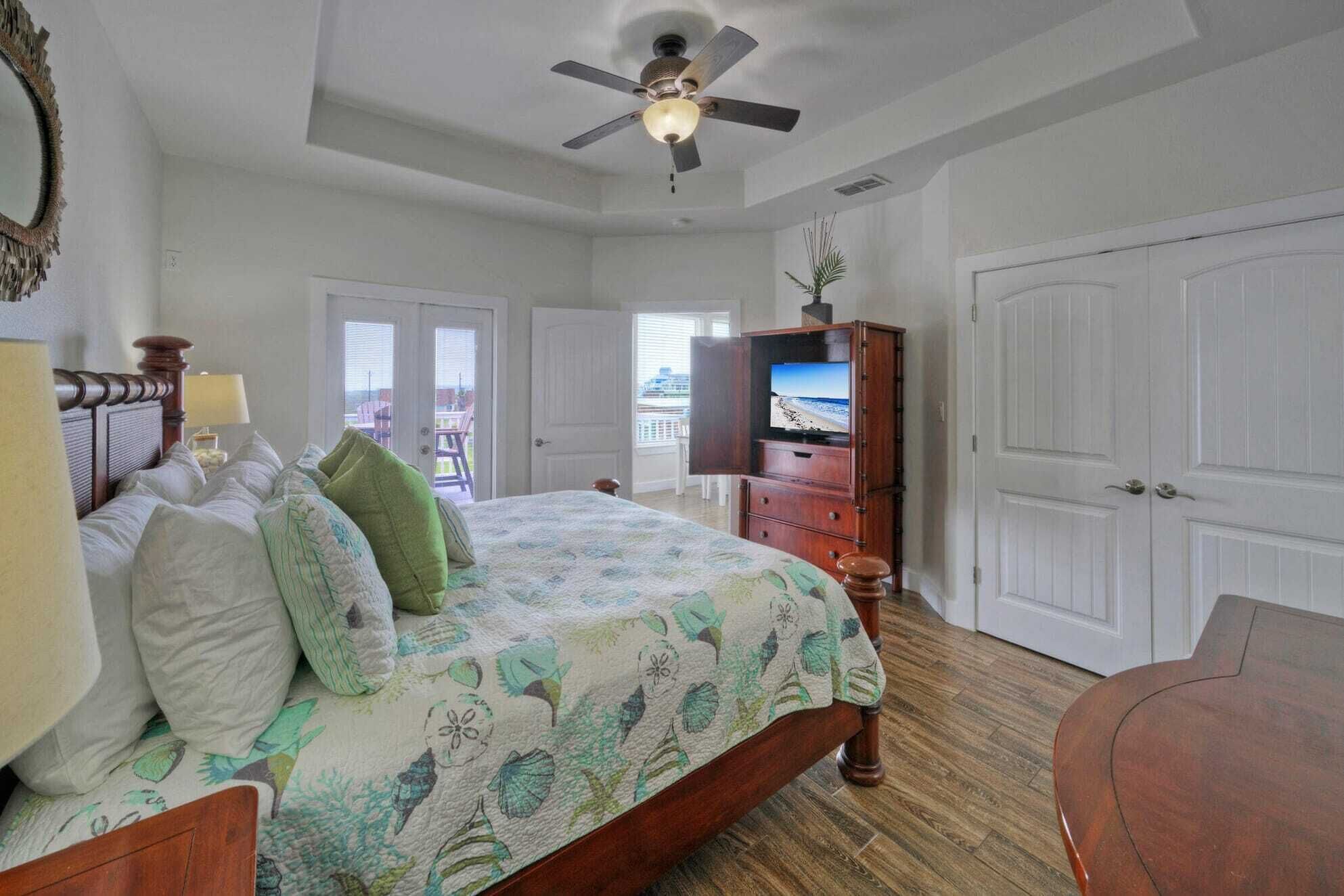A bedroom with a bed, a ceiling fan, a television, and a view of the beach through a door.