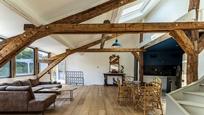 Um amplo loft com vigas de madeira aparentes, uma área de estar aconchegante com sofá e poltronas, uma mesa de jantar e uma cozinha com parede azul.