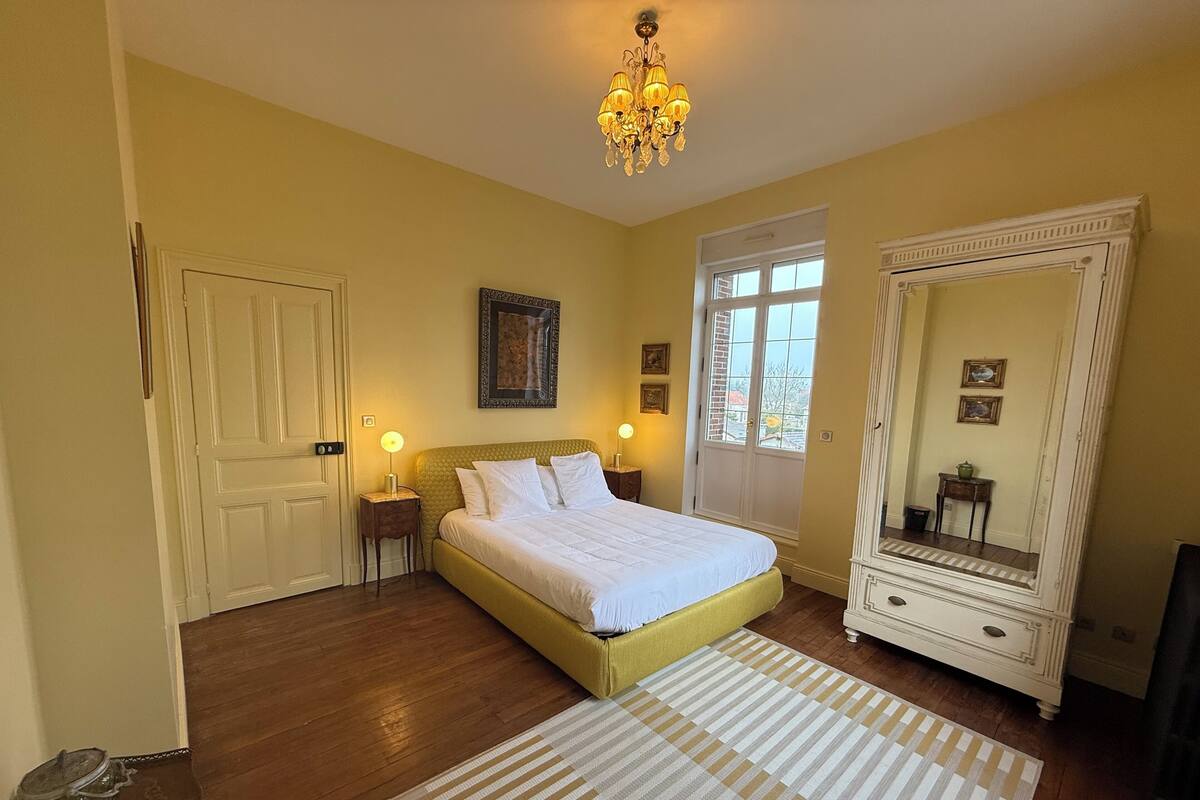 A bedroom with a large bed, a dresser, a mirror, a chandelier, and a door with a window.
