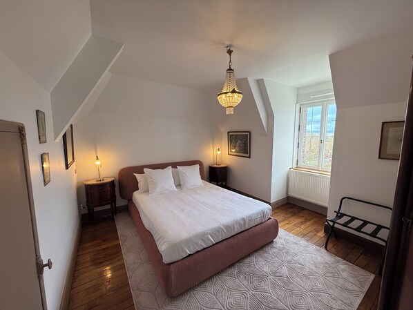 A bedroom with a large bed, a chandelier, a window with a view, and a radiator. - Château de Ligny (Ligny-le-Chatel)