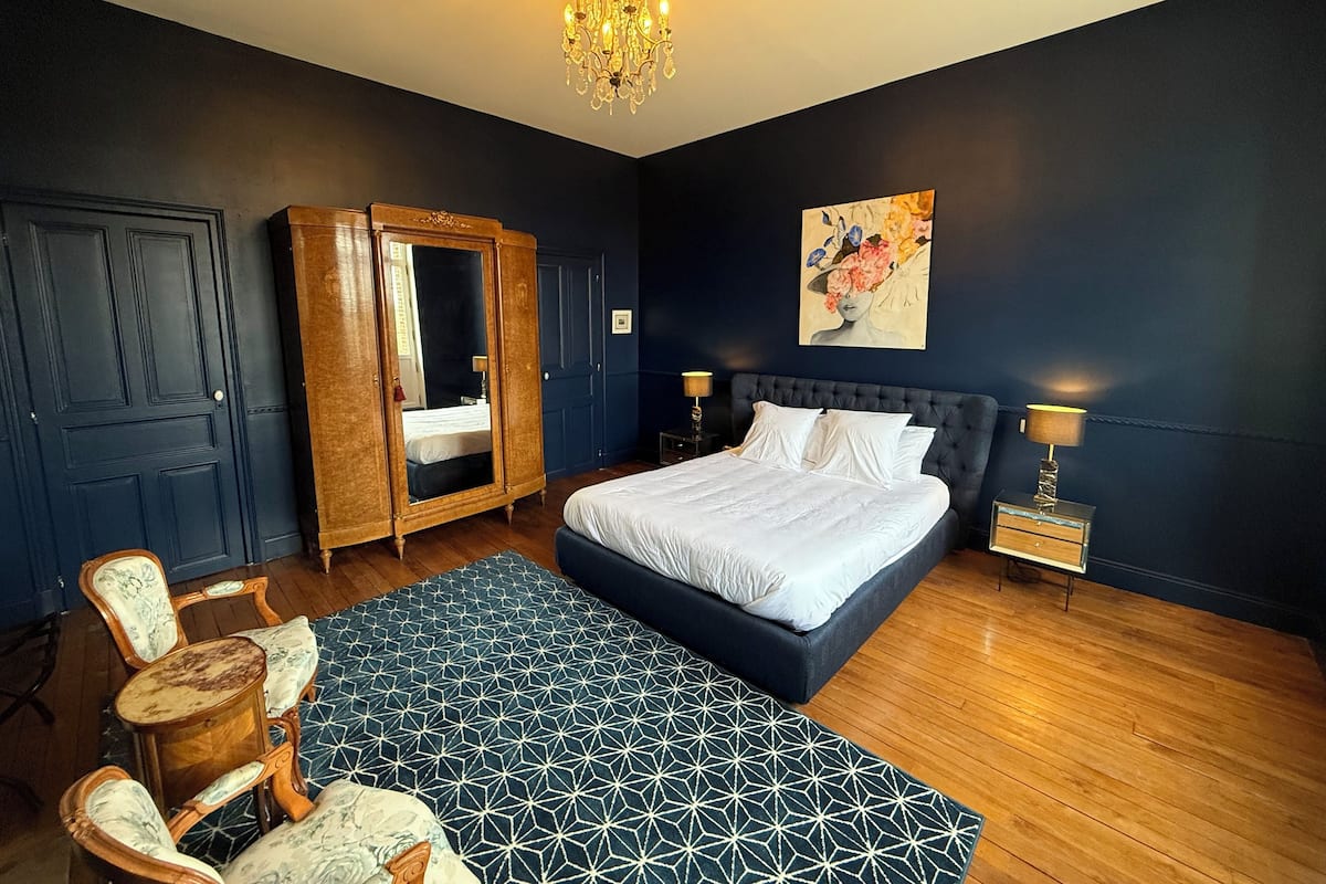 A bedroom with a large bed, a wardrobe, a mirror, a chandelier, and a painting on the wall.