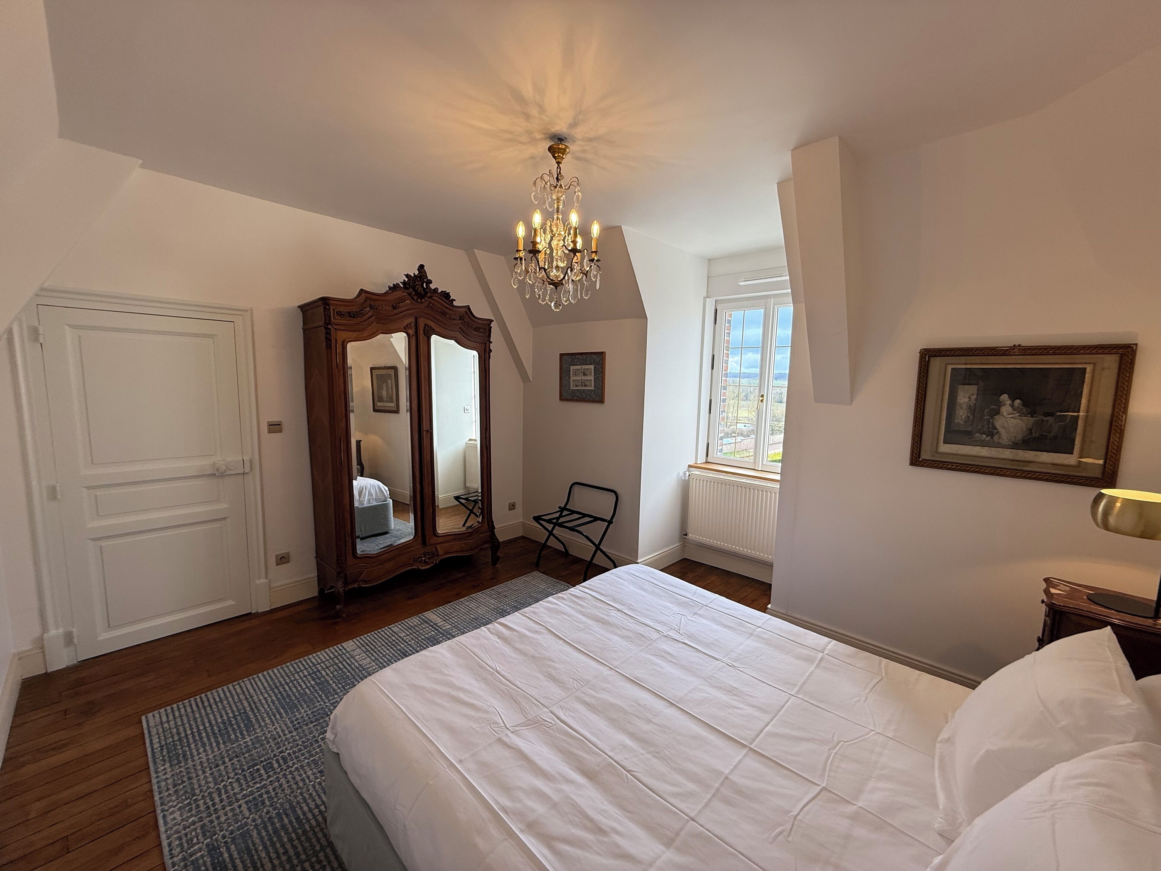 Un dormitorio con una cama grande, un armario de madera, una lámpara de araña, una ventana con vistas y un radiador.