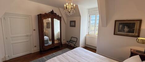 Un dormitorio con una cama grande, un armario de madera, una lámpara de araña, una ventana con vistas y un radiador.