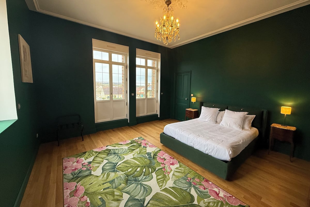 A bedroom with a large bed, two windows, a chandelier, and a floral rug.