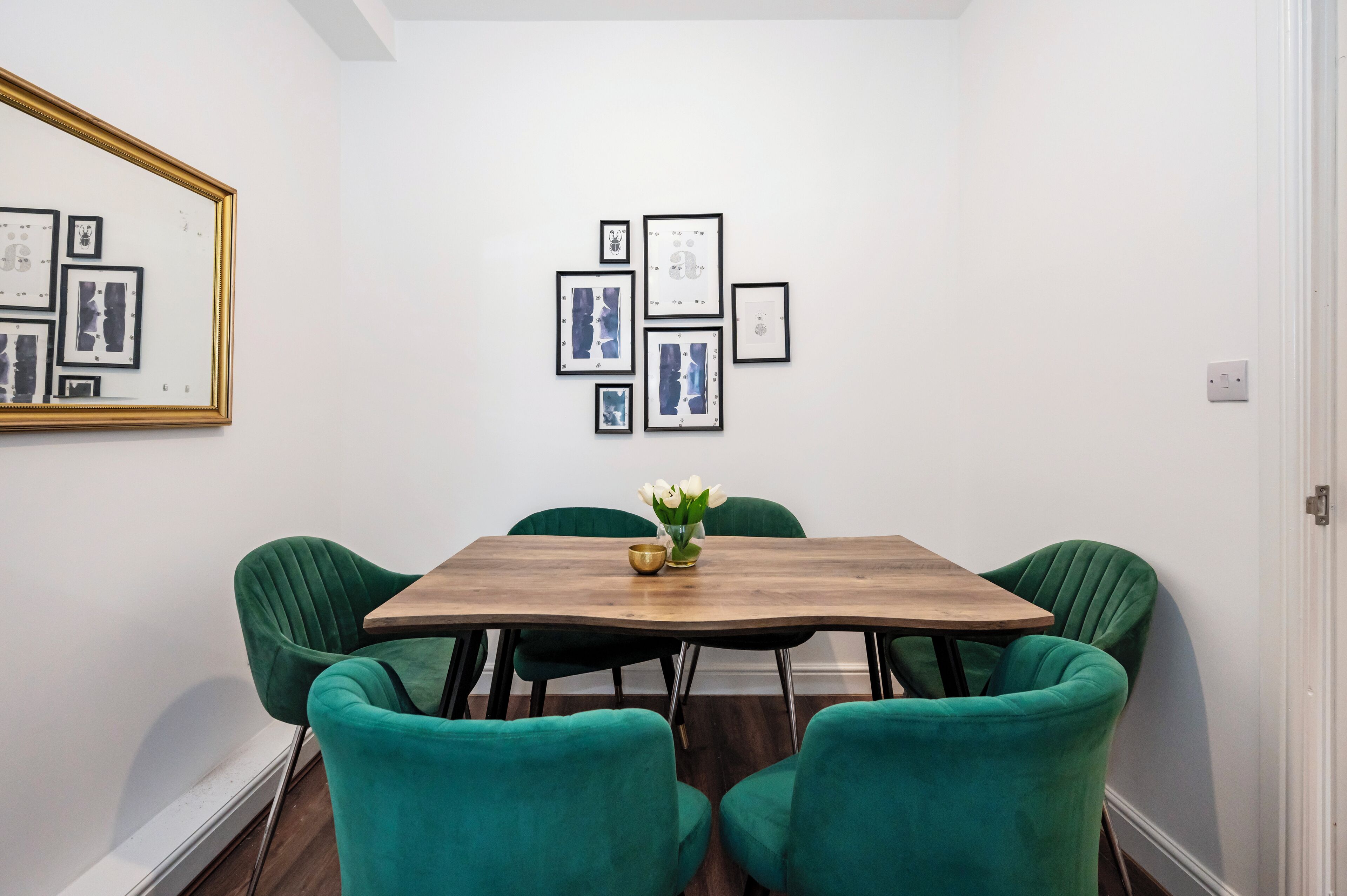 Een eethoek met een houten tafel en groene bekleden stoelen, kunstwerken aan de muur en een vaas met witte bloemen op tafel.