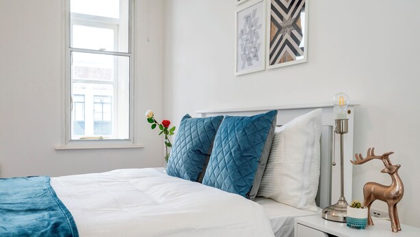 A neatly made bed with a blue blanket and pillows, a bedside table with a lamp, a decorative deer, and a window with a view. - Beautiful Flat Near the Center 11 min  (London)