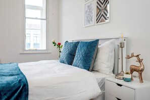 A neatly made bed with a blue blanket and pillows, a bedside table with a lamp, a decorative deer, and a window with a view. - Beautiful Flat Near the Center 11 min  (London)