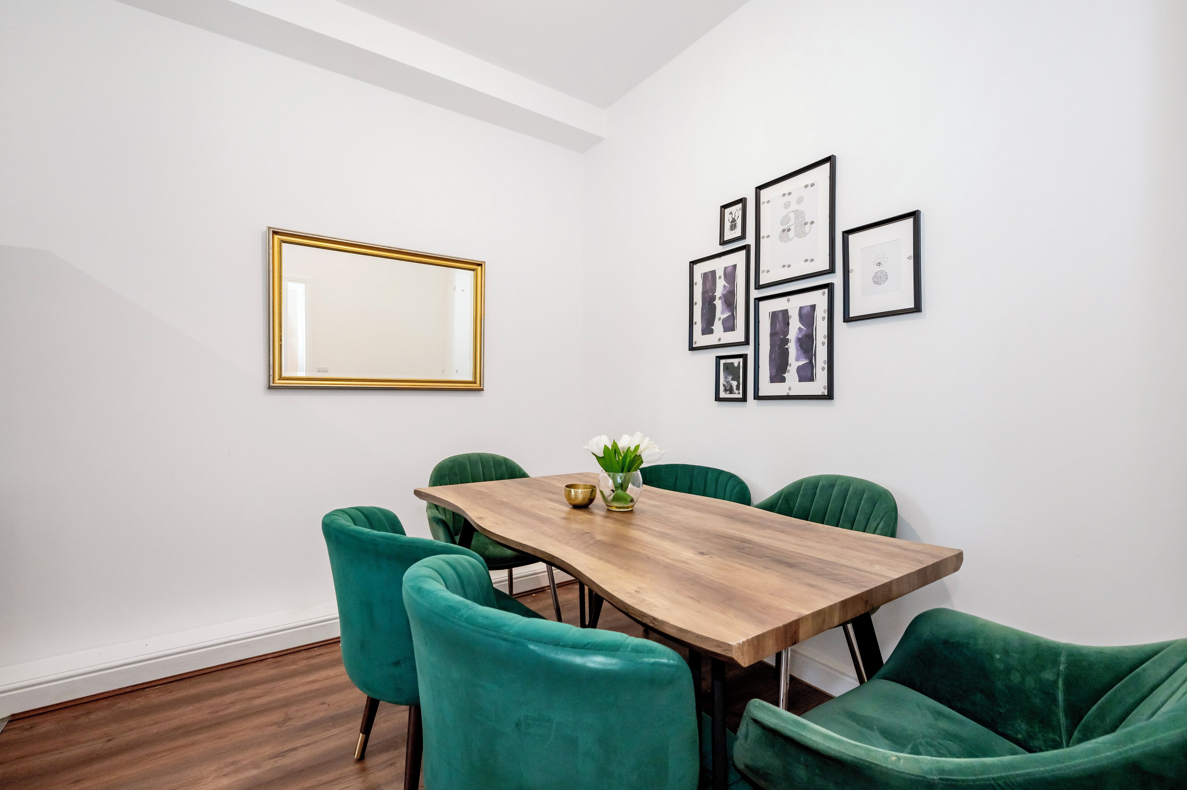 Un comedor con mesa de madera y sillas tapizadas en verde, un espejo con marco dorado y cuadros en la pared.