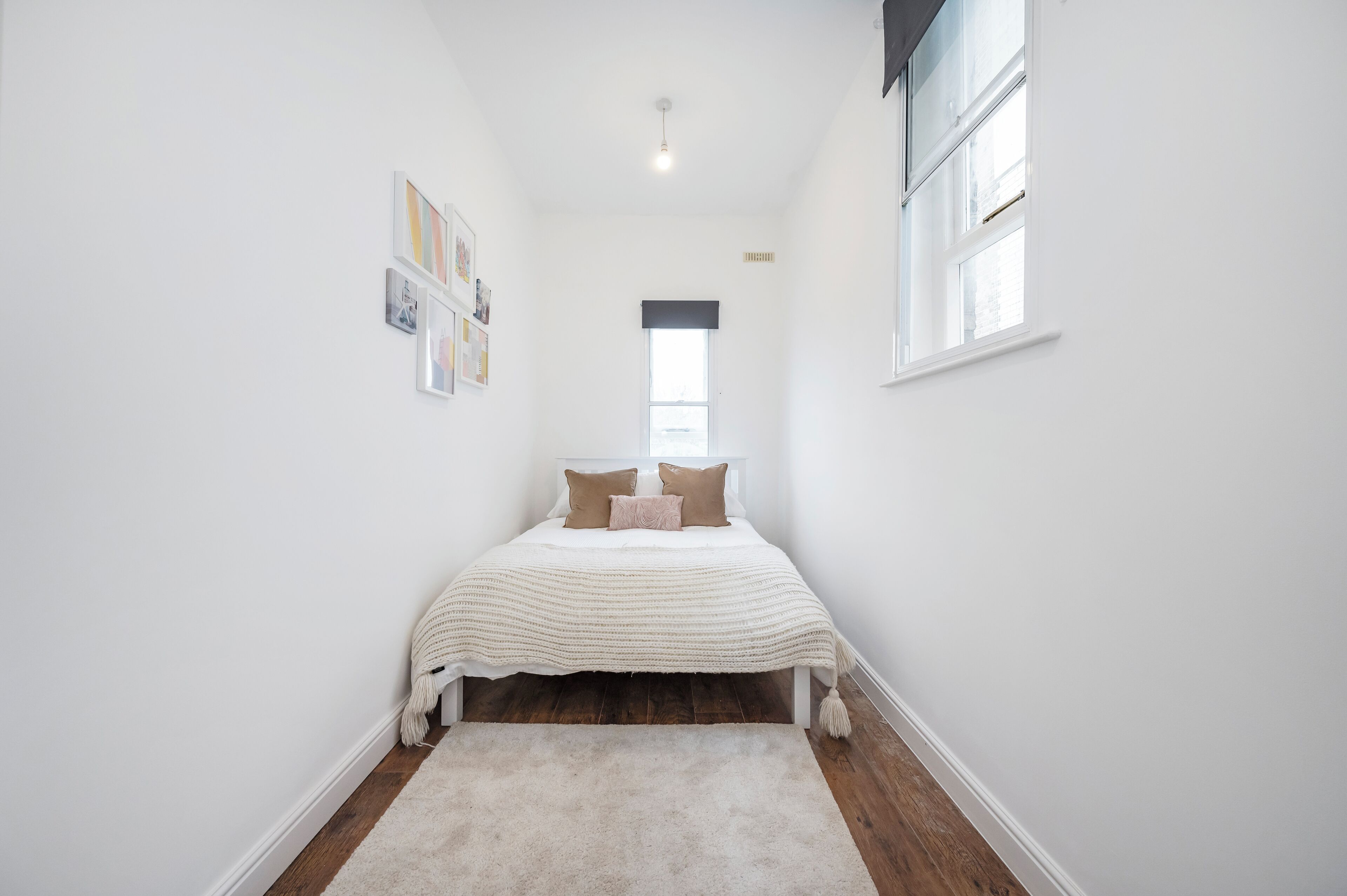 Un dormitorio estrecho con paredes blancas, una cama centrada debajo de una ventana, alfombra blanca y una lámpara de techo.