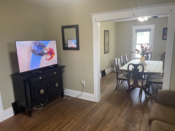 Un comedor con una mesa y sillas de madera, un televisor sobre un mueble negro y un pasillo que conduce a otra habitación.