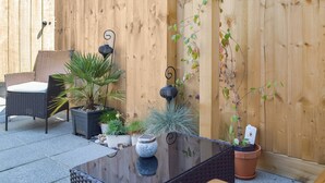 An outdoor patio with a wicker chair, a glass table, potted plants, and a wooden fence.