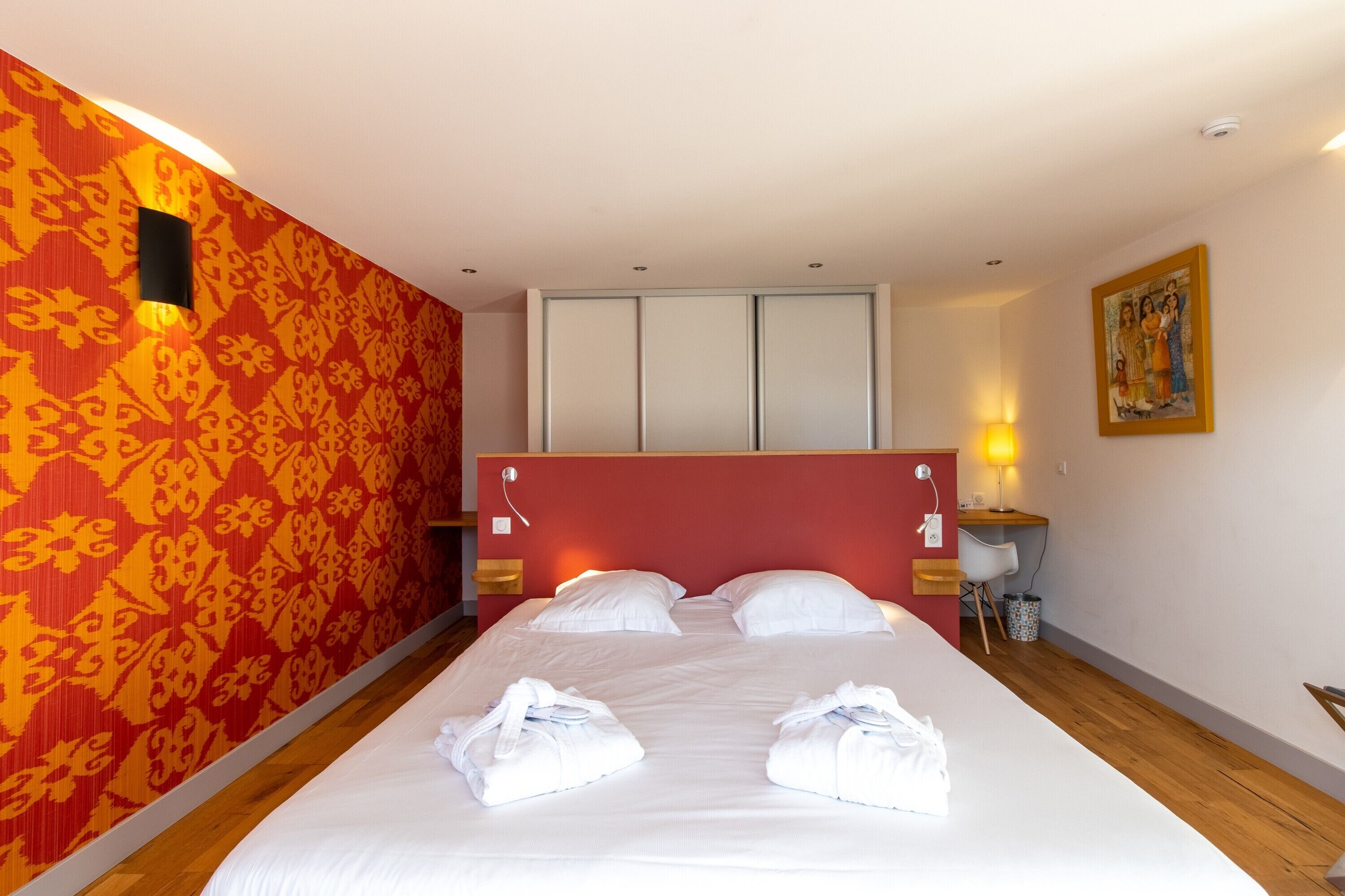 Una habitación de hotel con una cama, un cabecero rojo, un papel tapiz estampado, un cuadro y una mesita de noche con una lámpara.