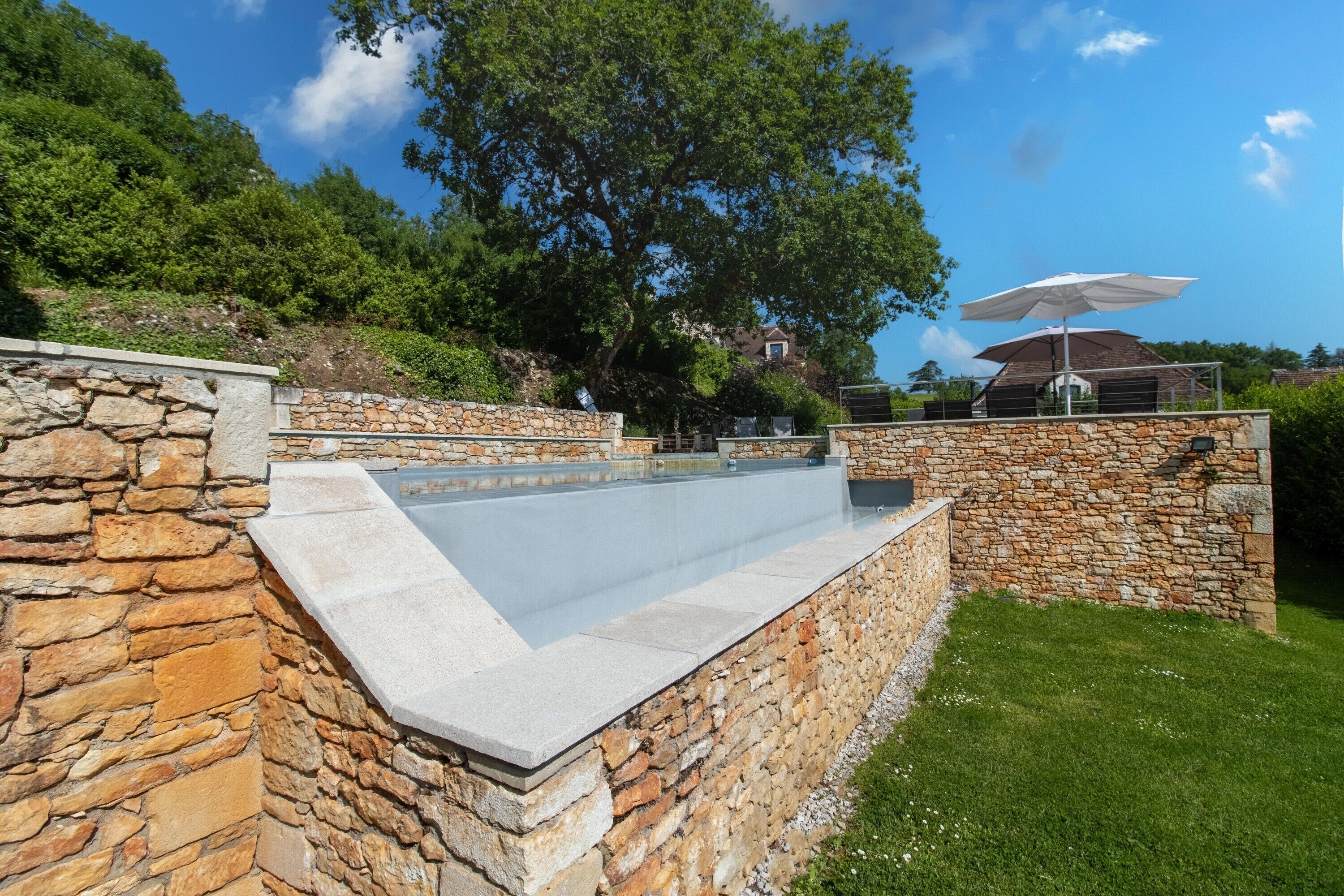 Una piscina con muros de piedra, un área con césped y una casa al fondo.