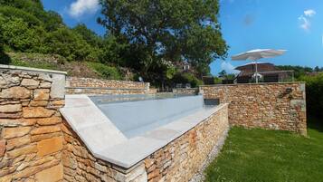 Una piscina con muros de piedra, un área con césped y una casa al fondo.