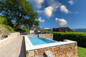 A swimming pool with stone walls, a wooden deck, and a view of houses and hills in the background.