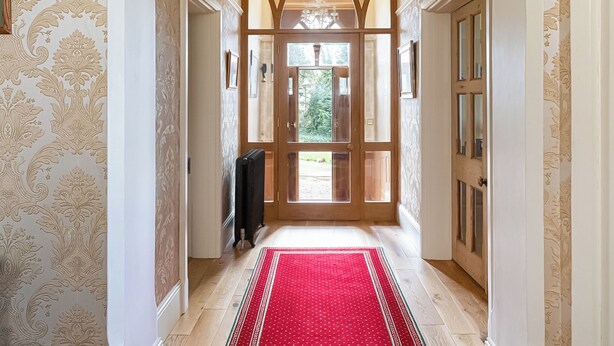 Un pasillo con papel tapiz estampado, alfombra roja, candelabro y puerta de madera que da al exterior.