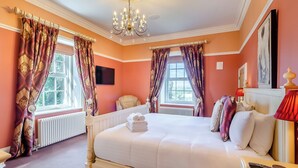 A spacious bedroom with a large bed, two windows with curtains, a chandelier, a radiator, a chair, and a bedside table with a remote control. - 6 bedroom accommodation in Old Quarrington, near Durham (Old Quarrington, near Durham)