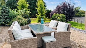 A wicker outdoor seating area with cushions and a glass-top table, surrounded by a garden with various plants and a wooden fence. - 6 bedroom accommodation in Old Quarrington, near Durham (Old Quarrington, near Durham)