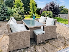 Un espace de détente extérieur aménagé avec des meubles en osier, doté de coussins et d’une table à plateau en verre, entouré d’un jardin où poussent différentes plantes et délimité par une clôture en bois.