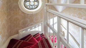 Una escalera con alfombra roja y barandilla blanca que conduce a una habitación con papel tapiz estampado y un ventanal circular.