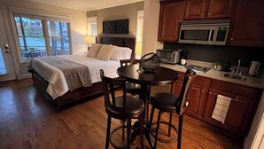 A bedroom with a bed, bedside tables, and a window. There is a kitchen area with wooden cabinets, a microwave, and a sink.