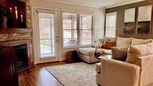 A living room with a fireplace, a beige sofa, a patterned rug, and a large window with blinds.
