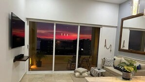 Un salon moderne doté d’une grande porte-fenêtre coulissante donnant sur un balcon, d’un téléviseur à écran plat fixé au mur, d’un canapé agrémenté de coussins décoratifs et d’une petite table sur laquelle est posée une plante.