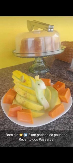 A plate of fruit arranged in the shape of a bird, with a cake in the background. - Pousada e Balneário Recanto dos Passaros BRB (Barreirinhas)