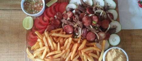 A wooden table with a variety of food items including sliced tomatoes, onions, meat, and French fries, along with small bowls of condiments.
