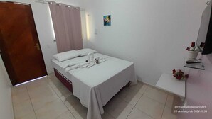 Ένα δωμάτιο με ένα μονό κρεβάτι, μια ξύλινη πόρτα, ένα λευκό κάλυμμα κρεβατιού, ένα μικρό τραπέζι με ένα βάζο με λουλούδια και έναν πίνακα που είναι κρεμασμένος στον τοίχο.