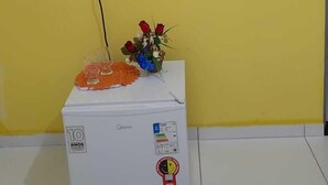 Ένα δωμάτιο με μια τηλεόραση επίπεδης οθόνης τοποθετημένη σε έναν κίτρινο τοίχο, ένα λευκό ψυγείο με ένα αυτοκόλλητο που υποδεικνύει την επέτειο των 10 ετών και ένα μικρό ράφι κάτω από την τηλεόραση.