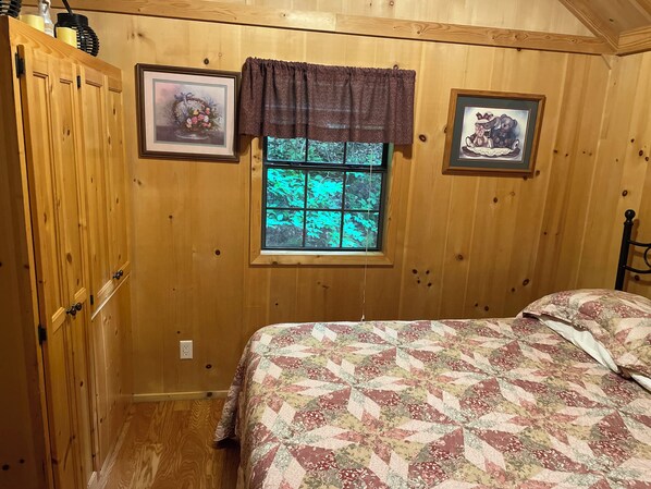 A bedroom with wooden walls, a window with a view of green foliage, a bed with a floral quilt, and two framed pictures on the wall. - Nature Retreat (South Bloomingville)