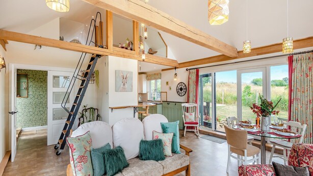 Ein großzügiger Wohnbereich mit hohen Decken, Holzbalken, einer Couch mit Zierkissen, einem Esstisch mit Stühlen und Blick ins Freie durch große Fenster.