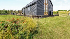 Eine dunkle Holzhütte mit einem großen Fenster, umgeben von einer grünen Wiese und roten Blumen.
