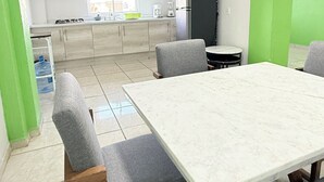 Un comedor con una mesa grande y cuatro sillas grises, una cocina al fondo con armarios y electrodomésticos, y una pared de acento verde.