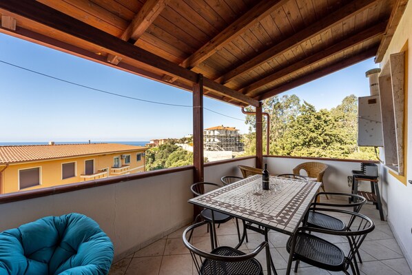 Varanda com teto de madeira, mesa e cadeiras azulejadas, com vista para uma área residencial e o mar.
