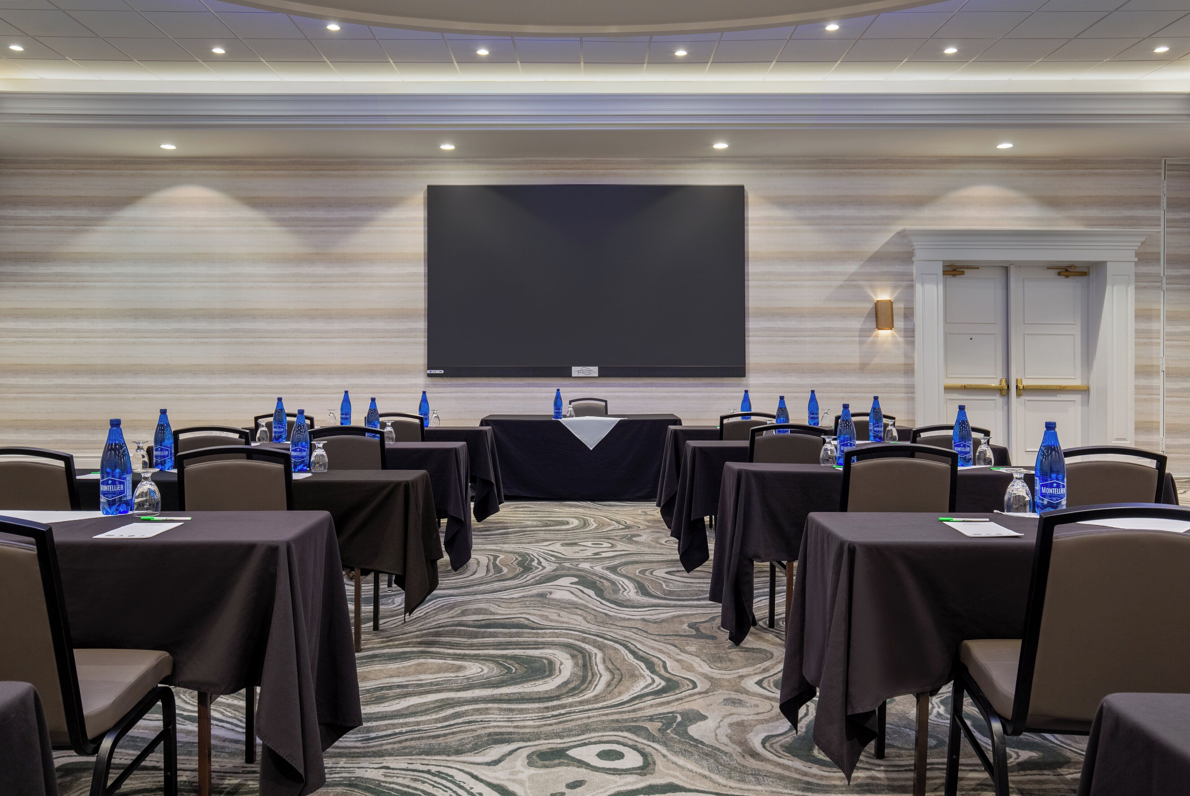 A conference room with a large screen, tables with chairs, water bottles, and a patterned carpet.