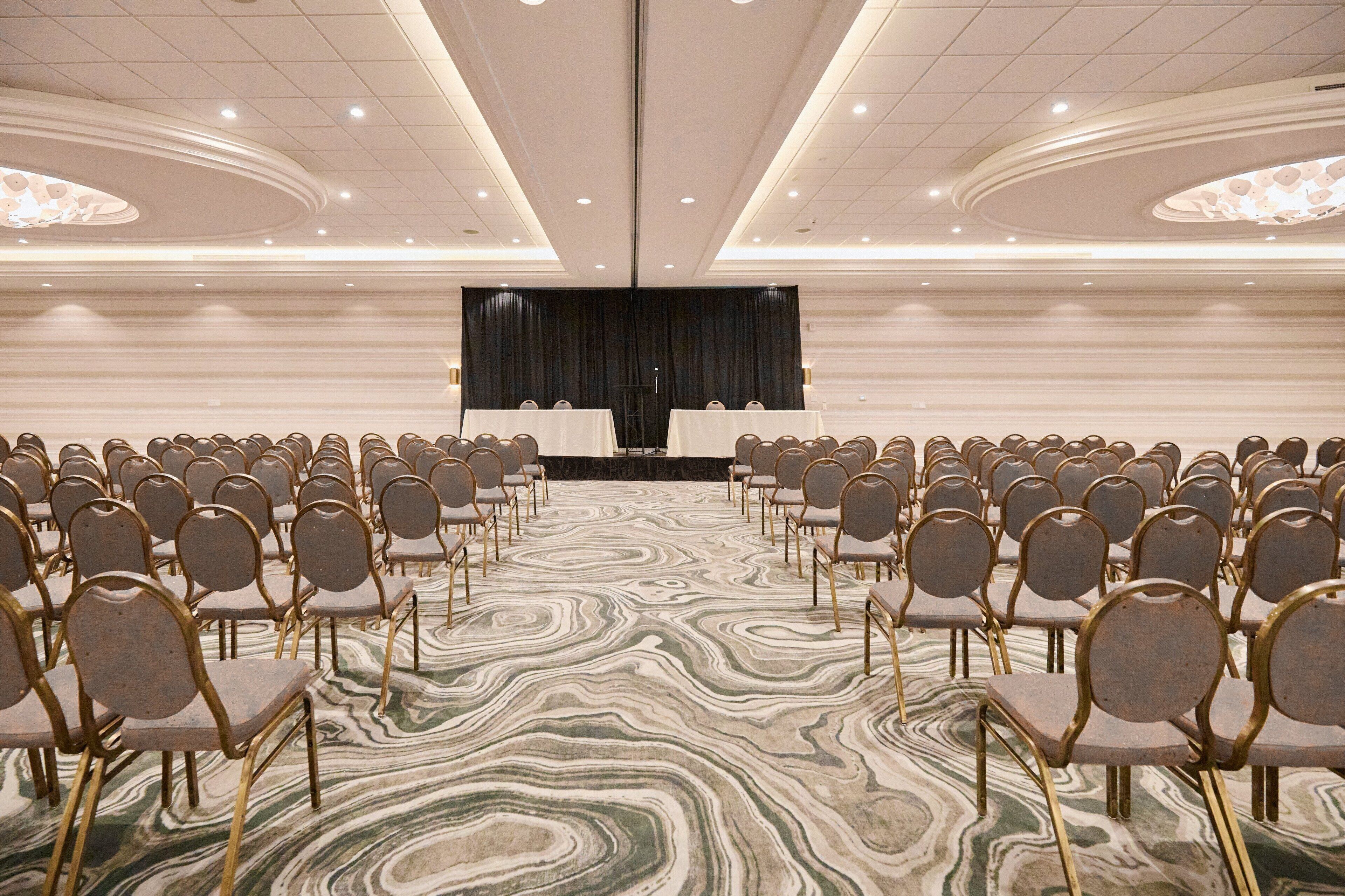 Un salón de conferencias con filas de sillas orientadas hacia un escenario, un suelo con diseño de mármol y un diseño moderno de techo.