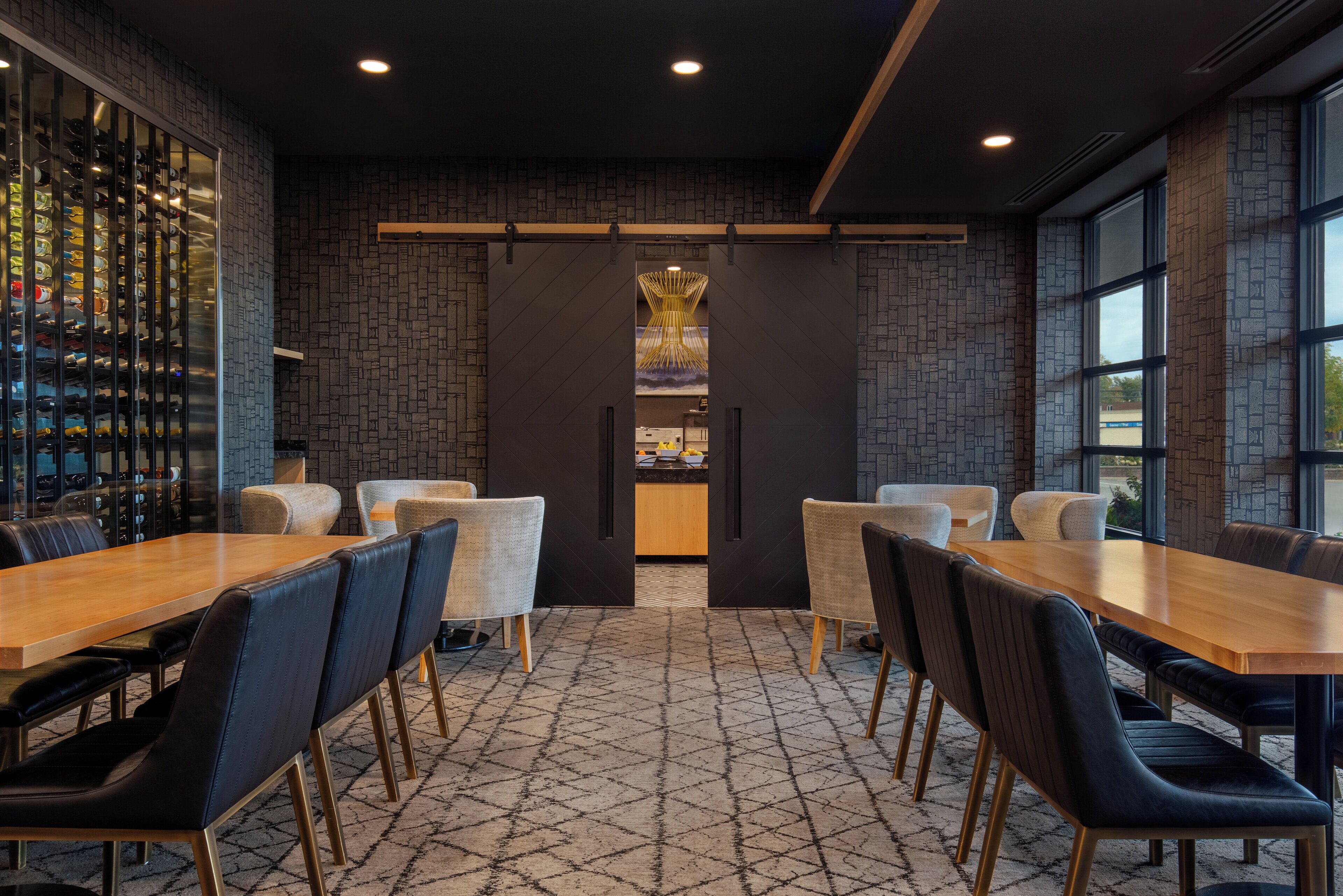 A modern dining area with a long wooden table and black upholstered chairs, a wine rack on the left, and a brick-patterned wall with a decorative light fixture above the counter.