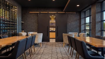 Un espace de salle à manger moderne avec une longue table en bois et des chaises à revêtement noir, un meuble à vin sur le côté gauche, et un mur décoré d’un motif en briques avec un luminaire décoratif au-dessus du comptoir.