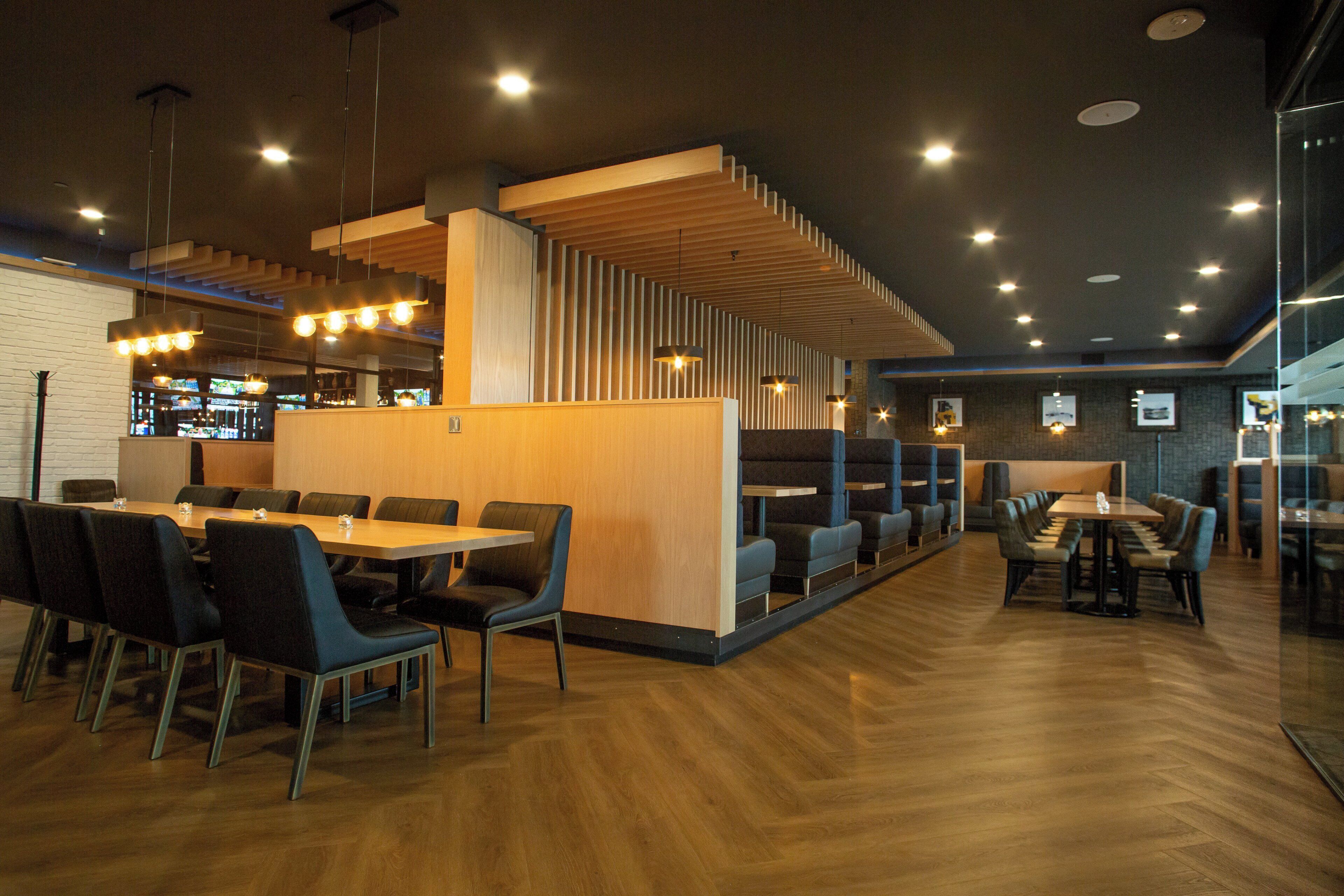 A modern restaurant with wooden tables and chairs, a bar area, and a seating section with cushioned benches.