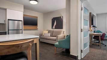 Una cocina moderna con refrigerador integrado, un comedor con mesa y sillas de madera, un sofá gris con cojines y un televisor de pantalla plana grande montado en la pared.