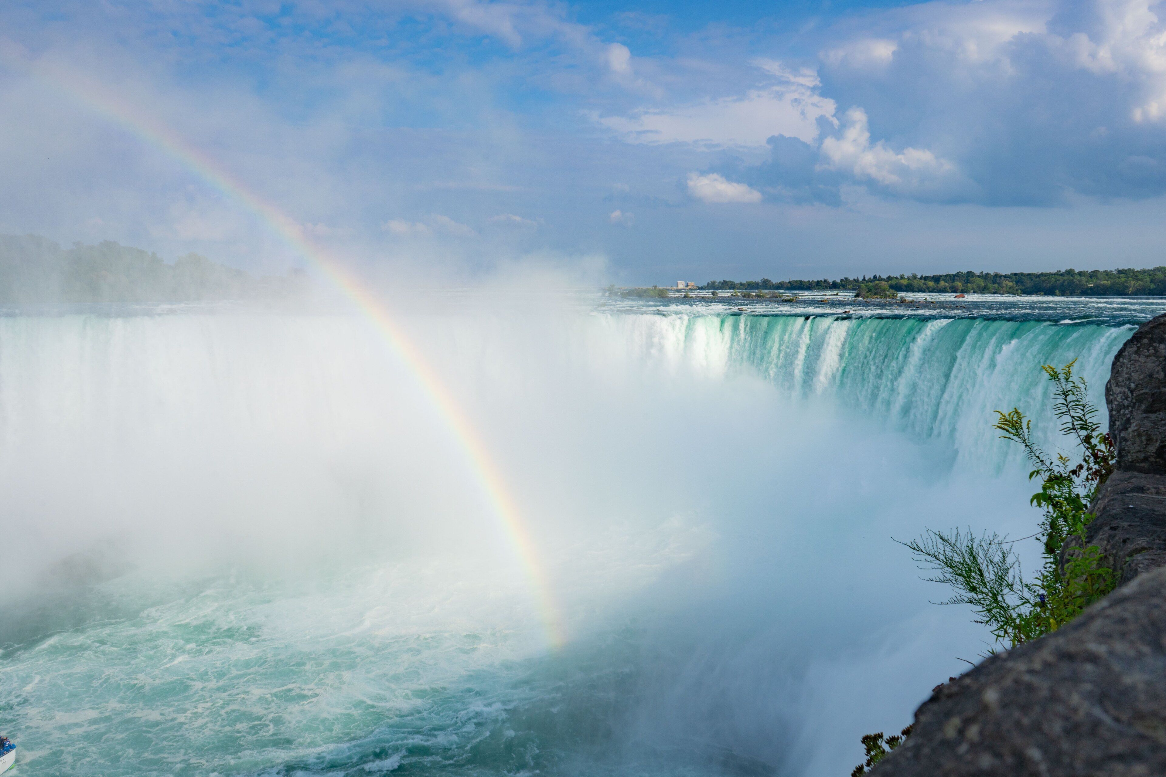 Ein Wasserfall mit Regenbogen, umgeben von felsigem Gelände und entfernten Gebäuden.