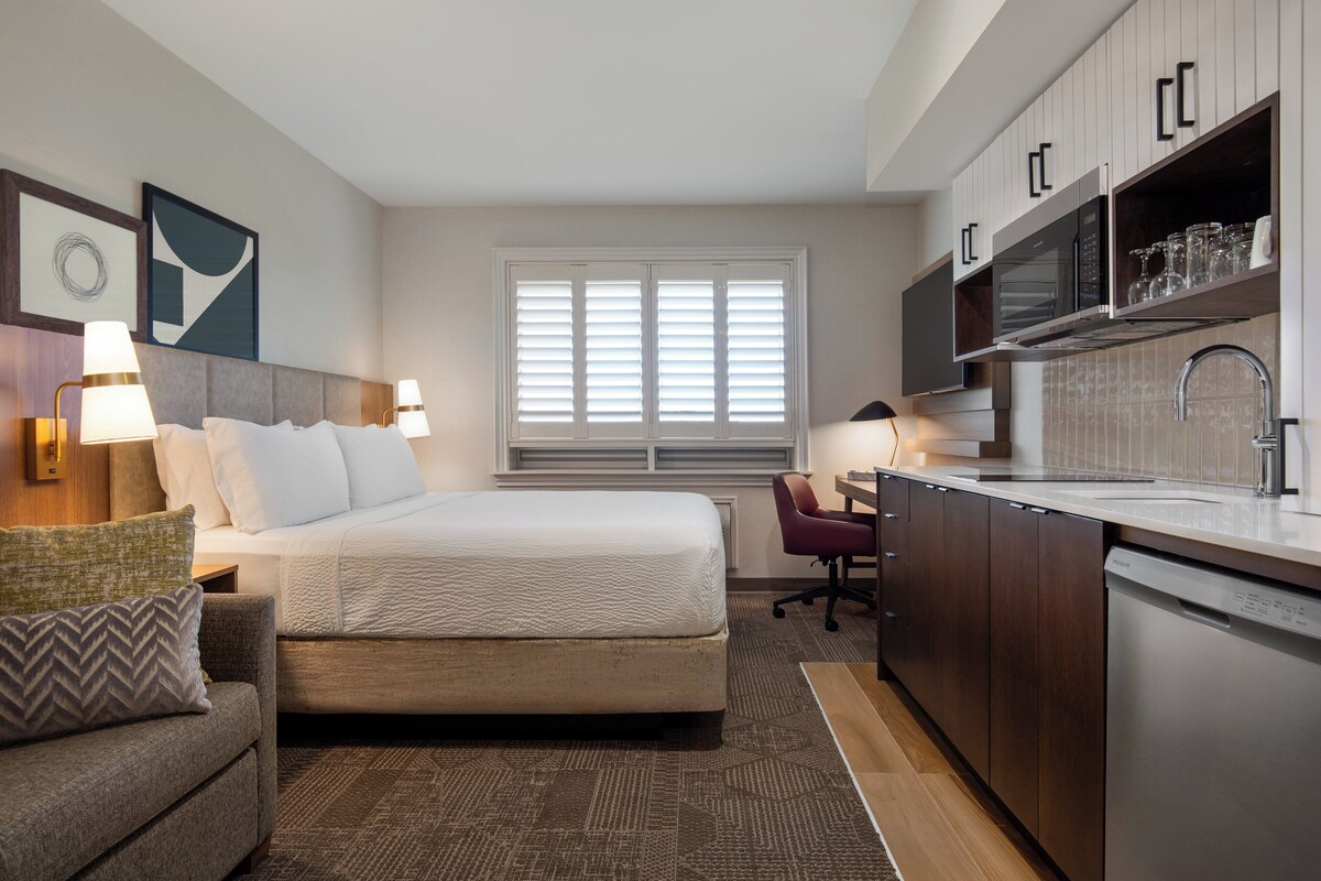 Een hotelkamer met een bed, een bureau, een stoel, een kitchenette en een raam met jaloezieën.