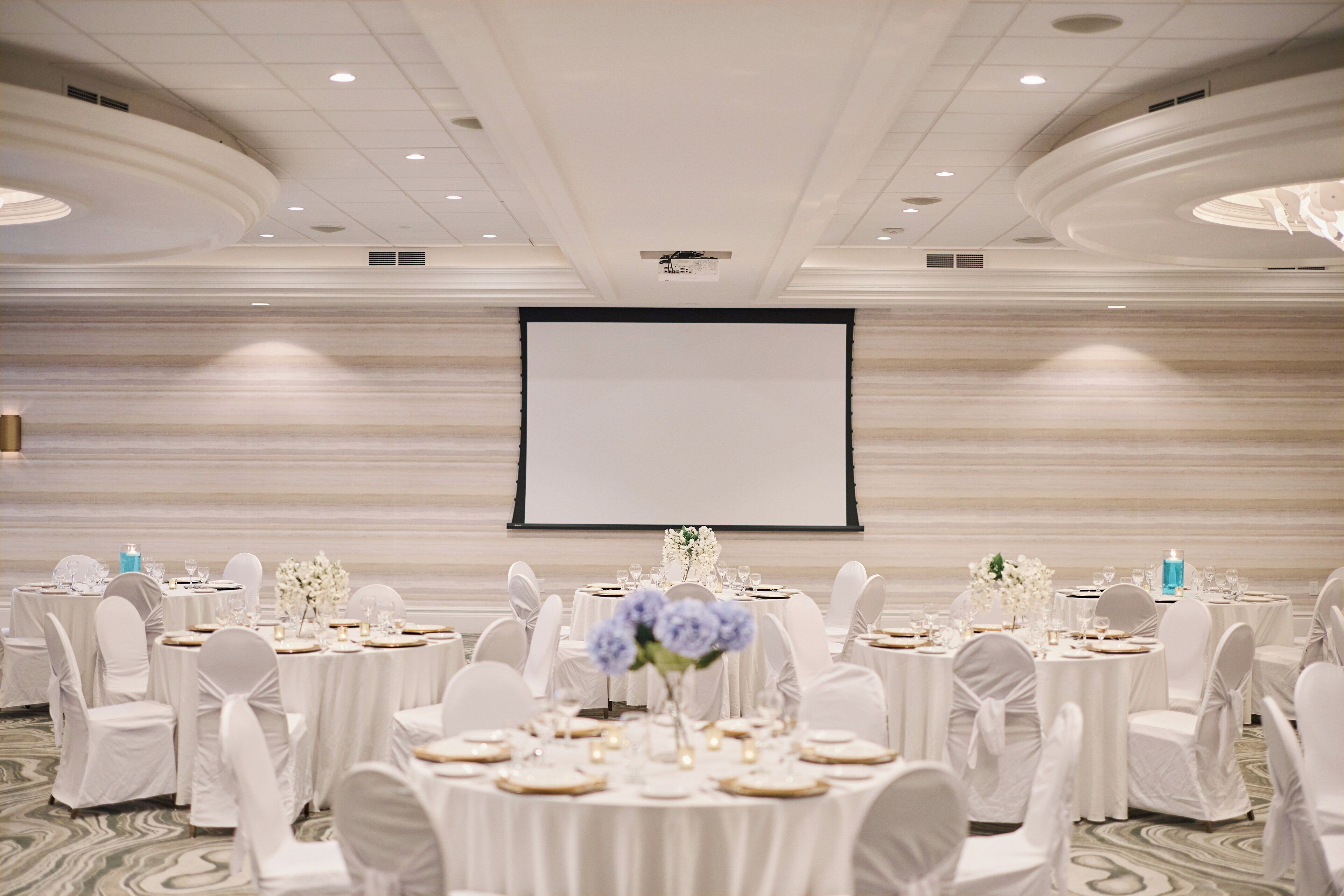 A banquet hall with round tables, white tablecloths, and chairs, a large screen, and a marble wall.