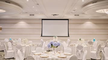 A banquet hall with round tables, white tablecloths, and chairs, a large screen, and a marble wall.