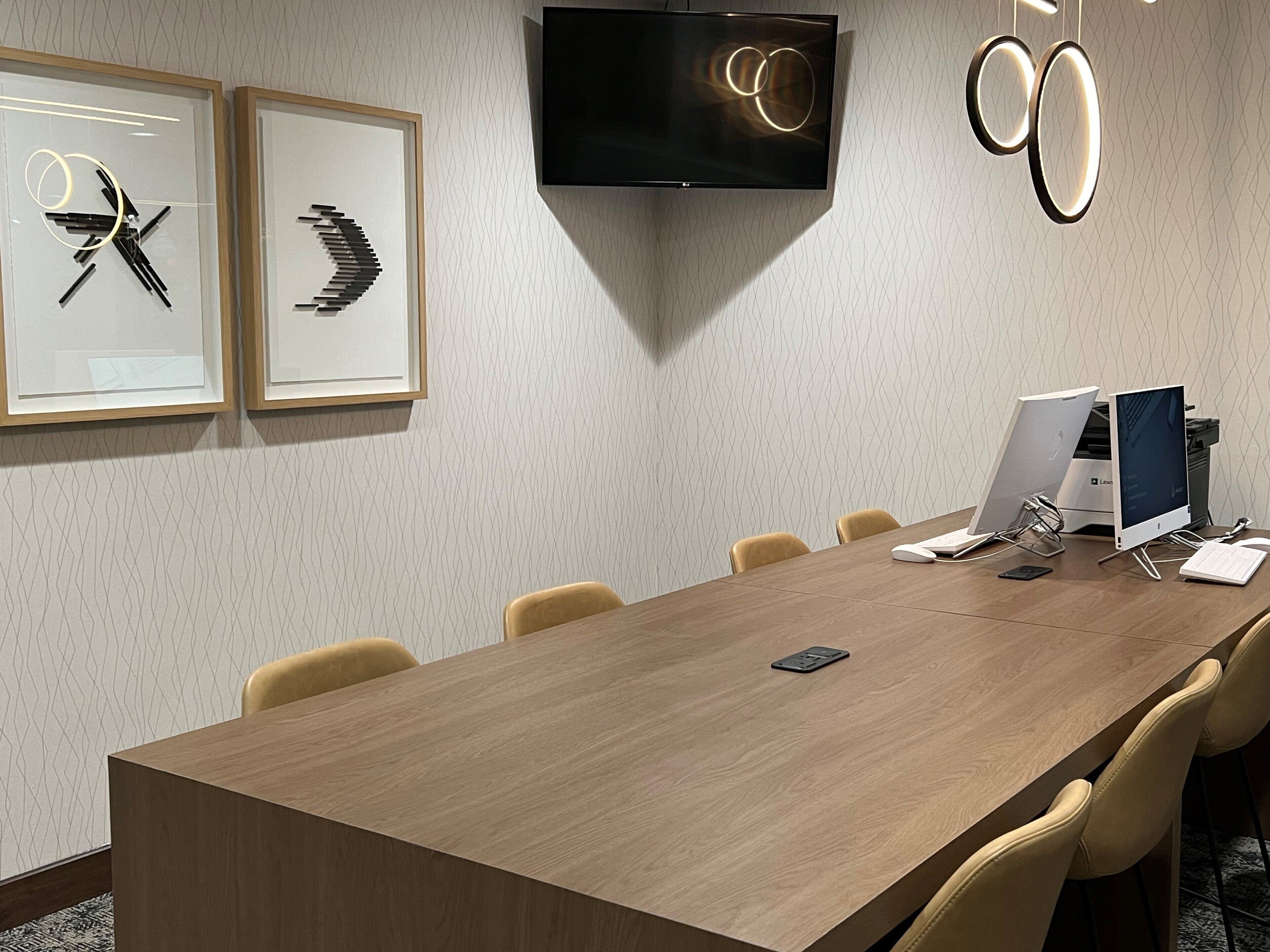 A conference room with a long wooden table, six chairs, a flat-screen TV mounted on the wall, and two framed abstract art pieces.