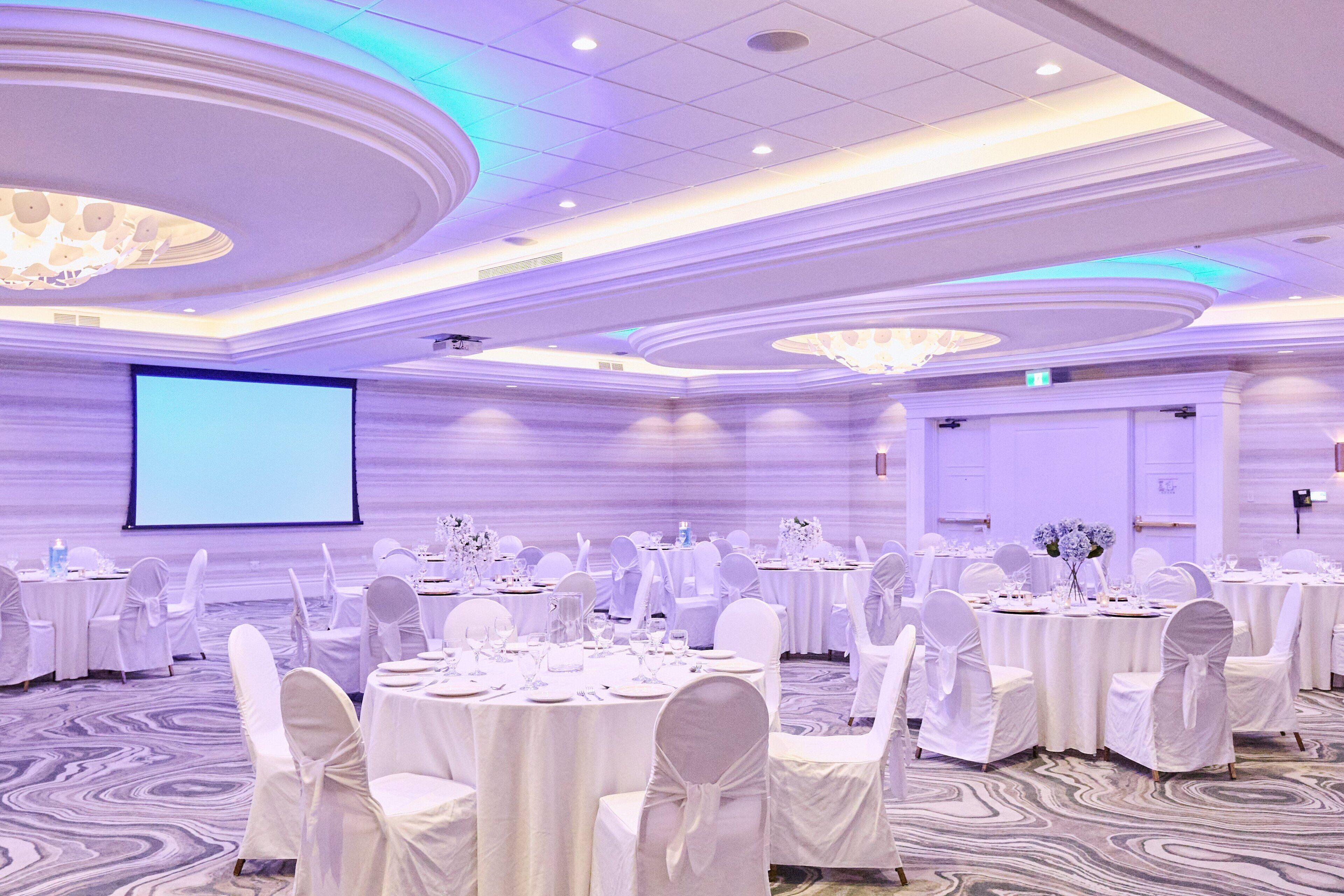 Une salle de banquet avec des tables rondes disposées pour les événements, des chaises blanches et un grand écran à l’avant.