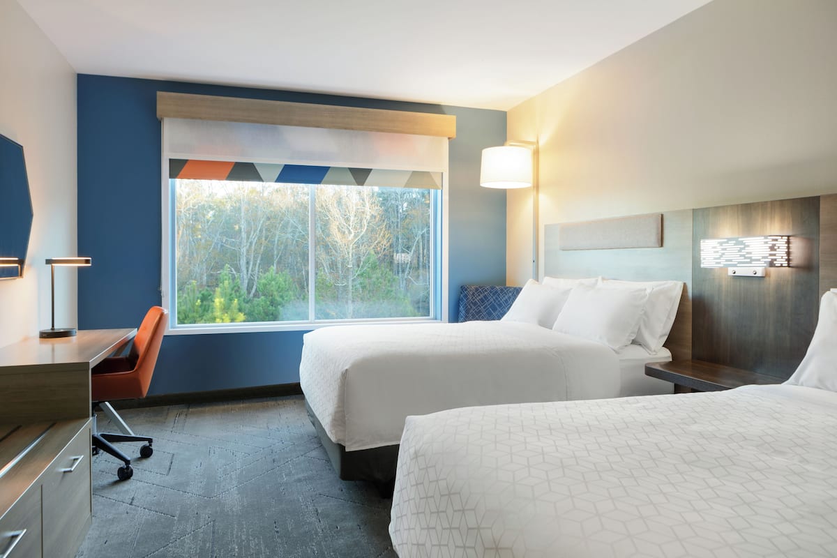 Ein Hotelzimmer mit zwei Betten, einem Schreibtisch, einem Stuhl und einem großen Fenster mit Blick auf Bäume.